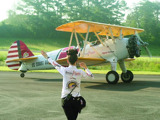 Dream Flights crew chief Jenny Garza waves a 1943 Boeing Stearman biplane carrying World War II veteran John Otwell and pilot Hunter Stuckey off the runway of the Gilmer County Airport. Three WWII veterans from Blue Ridge went up in the vintage plane last Tuesday.