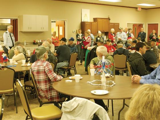 Sam Burrell, standing at left in light blue Air Force shirt, gives the good news to veterans, their spouses and others at the free breakfast on Veterans Day hosted annually by Ellijay First UMC. Early morning rain was slacking off, and the Lions Club parade and ceremony on the square would not have to be moved indoors.