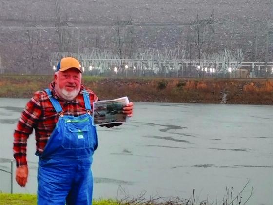Wayne Anderson stands in front of Carters Dam and the powerhouse during a 2024 interview. He’s holding a copy of a newsletter he subscribes to that did a historical section on the building of the dam in the 1960s-70s, showing photos of equipment and people he worked with. (Photo by Mark Millican)