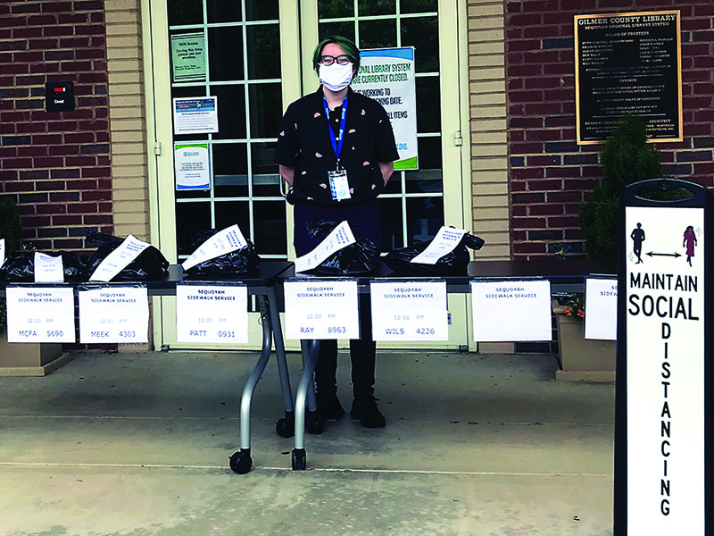 Gilmer County Library Sidewalk service is how the Gilmer County Library is safely checking out books and other material to customers. Above, staff member Cortny Crawford puts reserved material on the sidewalk service table in front of the local library.