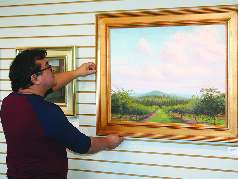 Jesus Morales, of the Gilmer Arts Gallery, straightens a painting by Atlanta artist Jill McGannon, whose work comprises one of three exhibits now on display at the downtown Ellijay gallery.