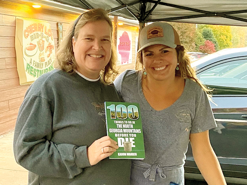 Karon Warren, left, holds a copy of her book, “100 Things to Do in the North Georgia Mountains Before You Die,” alongside Samantha Callihan, of The Pink Pig. The Cherry Log barbecue restaurant is among several Gilmer County businesses and attractions featured in the travel guide. 