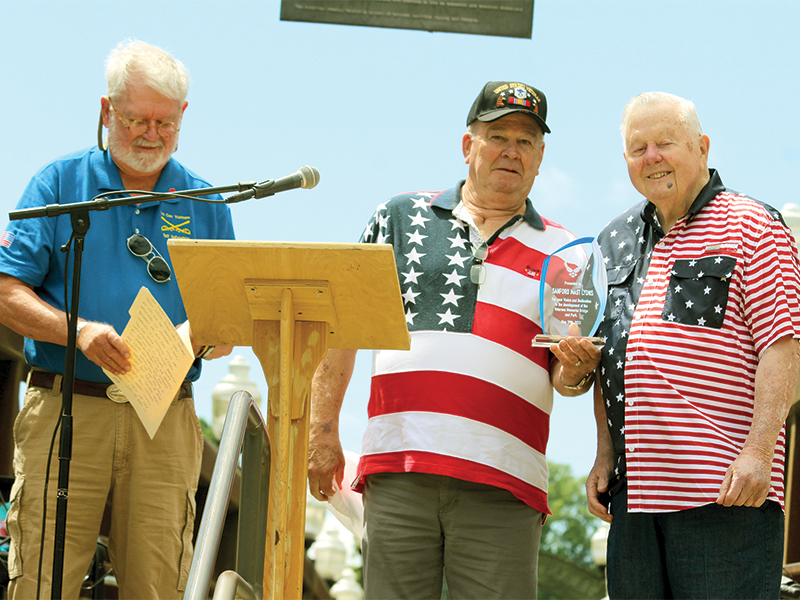Sandy Lyons, right, the driving force for the building of Veterans Memorial Bridge in the background, receives a Plaque of Appreciation from Don Murray of the Gilmer Veterans Memorial Committee on Memorial Day. At left, is committee member Ben Arp, who noted Lyons’ “tireless efforts” during the project that began in 1997 and was dedicated in 2009.